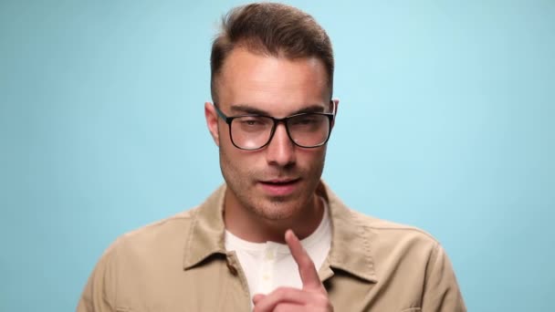 young casual man wearing jacket and eyeglasses and asking for silence on blue background - Footage, Video