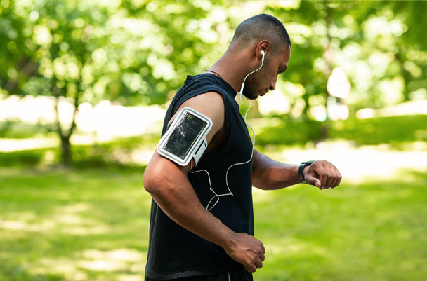 Side view of African American guy checking health data on smartwatch after jogging at park - Photo, Image