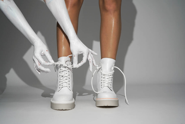   kobieta wiąże sznurowadła na białych butach na jasnym tle w studio. zbliżenie moda obuwie nowa kolekcja jesień zima 21 / 22 moda strzelanie                             - Zdjęcie, obraz