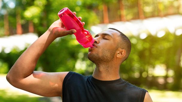 Жаждущий черный мужчина пьет воду из спортивной бутылки после тренировки в парке - Фото, изображение