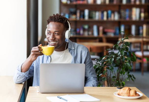 Chico negro feliz con auriculares escuchando música y tomando café aromático en la acogedora cafetería, espacio vacío - Foto, Imagen