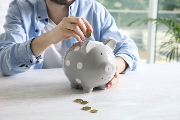 Man putting coin into piggy bank at white table indoors, closeup - Foto, Imagem