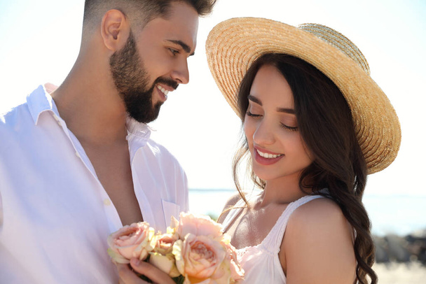 Happy young couple with flowers at beach near sea. Honeymoon trip - Zdjęcie, obraz