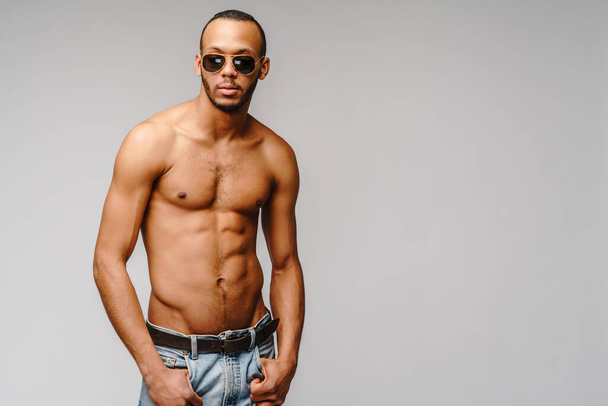 Sexy junge muskulöse afrikanisch-amerikanische Mann hemdlos trägt Sonnenbrille über hellgrauem Hintergrund - Foto, Bild
