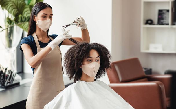 Hairdresser cutting hair of african woman, both wearing protective masks - Photo, Image