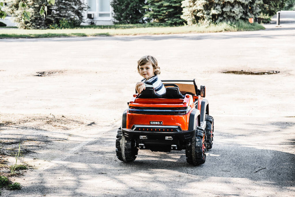 menino de cabelos encaracolados em uma camiseta listrada monta um carro de brinquedo vermelho grande dirigindo em um caminho de asfalto. dia de folga, recreação ao ar livre
 - Foto, Imagem