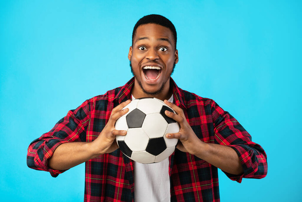 Emotionele zwarte man schreeuwen vasthouden voetbal bal op blauwe achtergrond - Foto, afbeelding