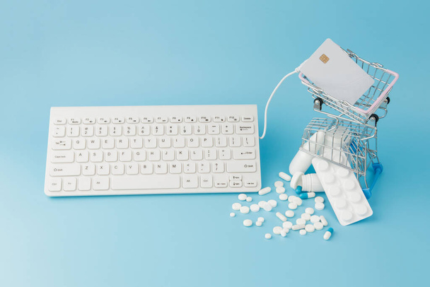 Shopping cart toy with medicaments and Keyboard. Pills, blister packs, medical bottles, thermometer, protective mask on a blue background. . Health care and internet shopping. Top view with place for your text. - 写真・画像