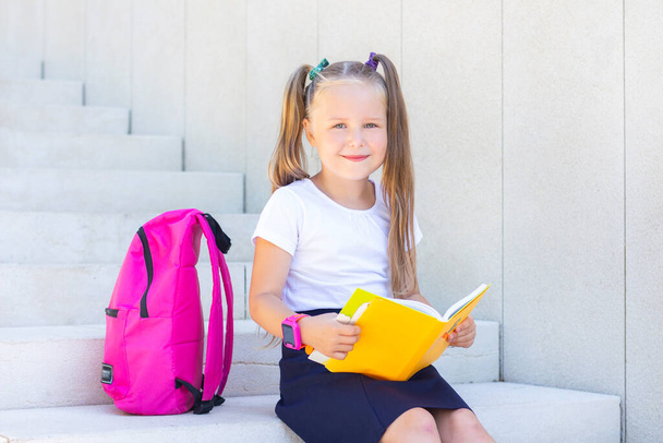 schoolgirl sits with a backpack and a textbook and smiles. - Foto, afbeelding