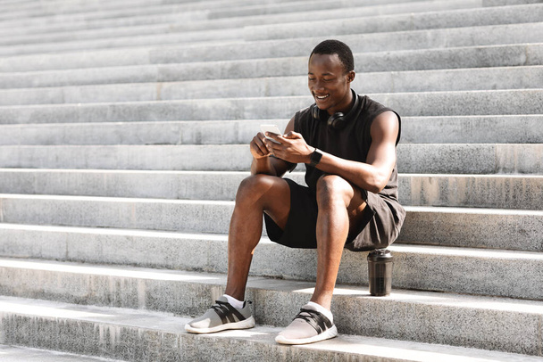 Deportivo chico negro usando smartphone después de entrenar al aire libre, sentado en escaleras urbanas - Foto, Imagen
