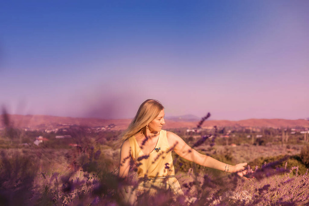 Blonde woman in a yellow dress in lavender field at sunset. Lavender bloom season. Enjoyment of unity with nature. Meditation and relaxation outdoor - Photo, image