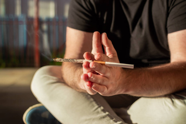 Junger Mann sitzt mit Cannabis-Joint und Zigarette auf der Straße. - Foto, Bild