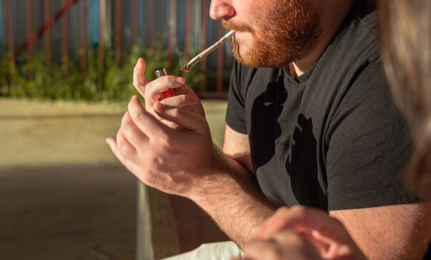 Young man with red beard smoking weed joint on the street at sunset. - Photo, image