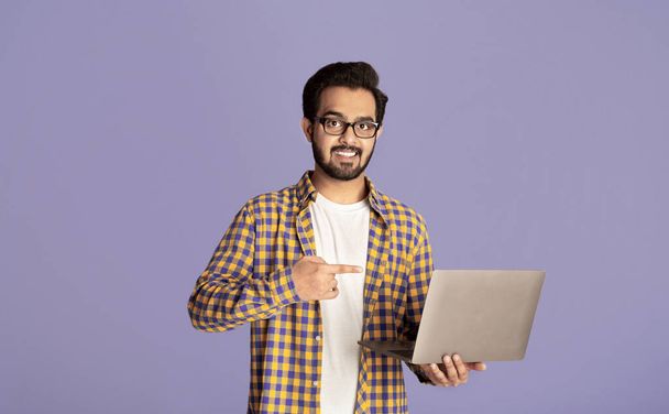 Ηλεκτρονική εργασία, εκπαίδευση, επικοινωνία. Ευτυχισμένος Ινδός που δείχνει το λάπτοπ πάνω από το λιλά φόντο. - Φωτογραφία, εικόνα