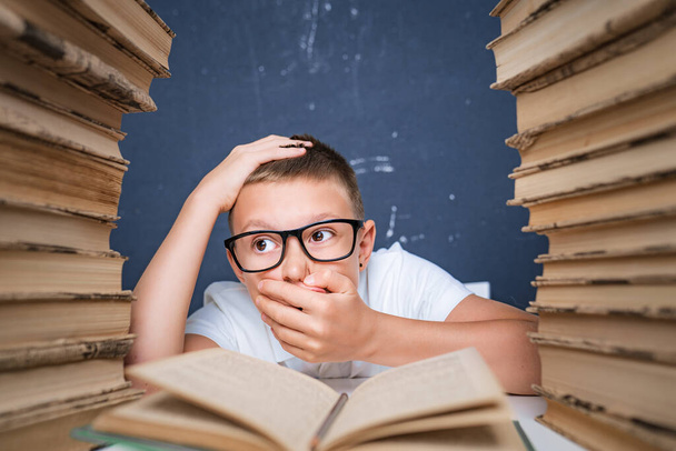 Smart boy in glasses getting crazy of reading book while sitting between two piles of books - Zdjęcie, obraz