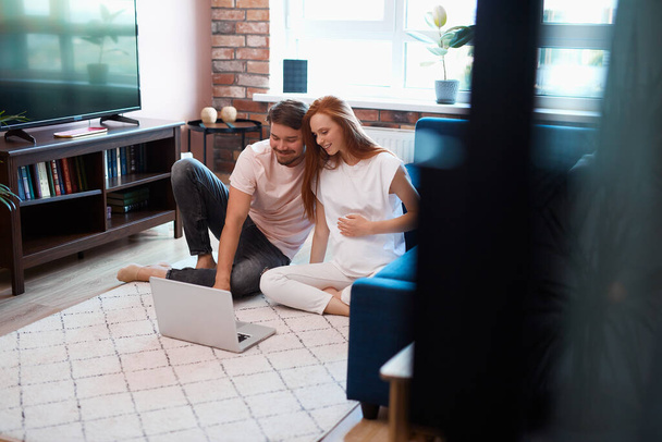 adorable future parents sit on the floor with laptop - Фото, изображение