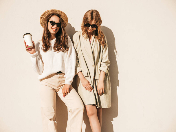 Two young beautiful smiling hipster girls in trendy white sweater and coat. Sexy carefree women posing near wall in the street in hat and sunglasses. Positive models having fun and drinking coffee  - Фото, изображение