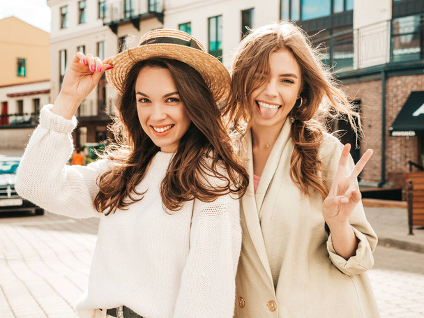 Two young beautiful smiling hipster girls in trendy white sweater and coat. Sexy carefree women posing on street background in hat. Positive models having fun. They show peace sign - Фото, изображение