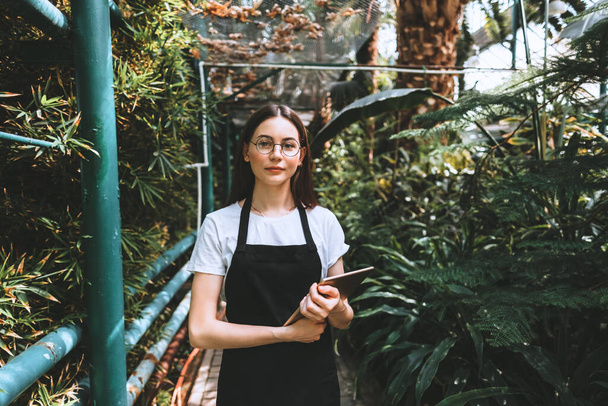 Portrait of young woman gardener in glasses and apron with digital tablet working in a garden center, looking at camera. Environmentalist using digital tablet in greenhouse. High quality photo - Photo, image