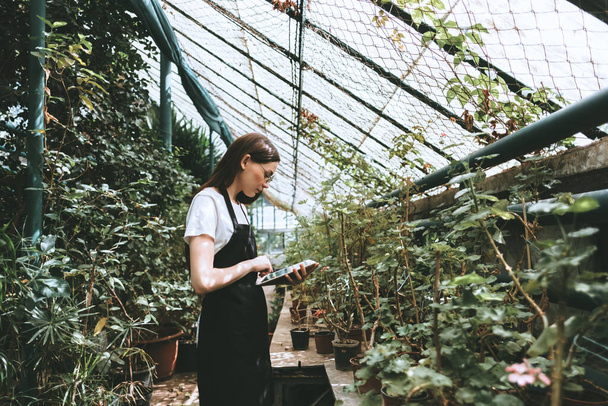 Young woman gardener in glasses and apron with digital tablet working in a garden center for better quality control. Environmentalist using digital tablet in greenhouse. High quality photo - Fotó, kép