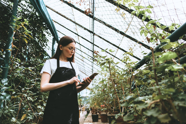 Young woman gardener in glasses and apron with digital tablet working in a garden center for better quality control. Environmentalist using digital tablet in greenhouse. High quality photo - Photo, Image