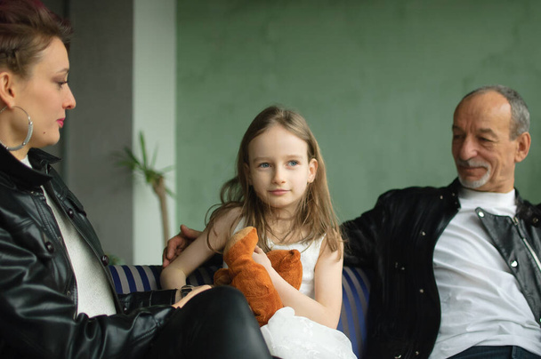 Familienporträt der erwachsenen Tochter, der kleinen Enkelin und des älteren Großvaters im Dachgeschoss mit Zimmerpflanzen. Mann und Frau tragen schwarze Lederjacken im Punk-Stil, Generationen-Konzept - Foto, Bild