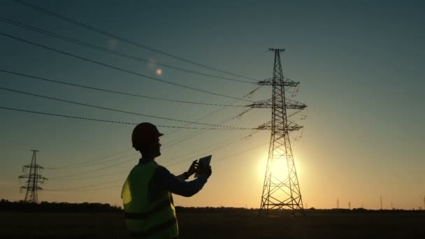 Pracovník v uniformě kontroluje elektrické vedení pomocí tabletu na pozadí západu slunce. - Záběry, video