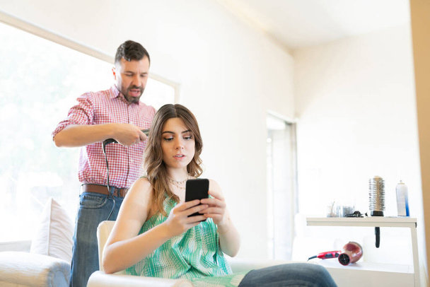 Junge Frau benutzt Mobiltelefon, während der lateinische männliche Friseur ihr Haar frisiert - Foto, Bild