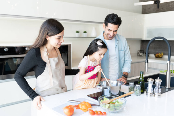 Улыбающаяся латинская семья смотрит на дочь, готовящую еду на кухне - Фото, изображение