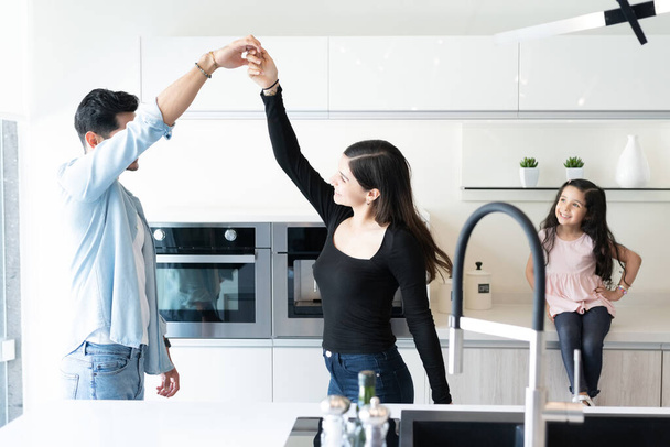 Happy daughter sitting on countertop and looking at parents dancing in kitchen - Foto, Imagem