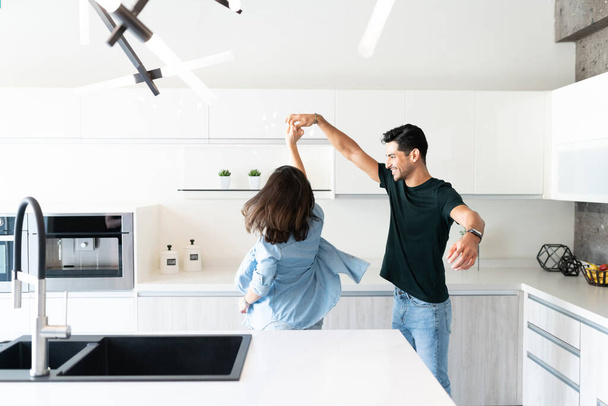 Χαμογελώντας όμορφος νεαρός που χορεύει με την κοπέλα του στην κουζίνα στο σπίτι - Φωτογραφία, εικόνα