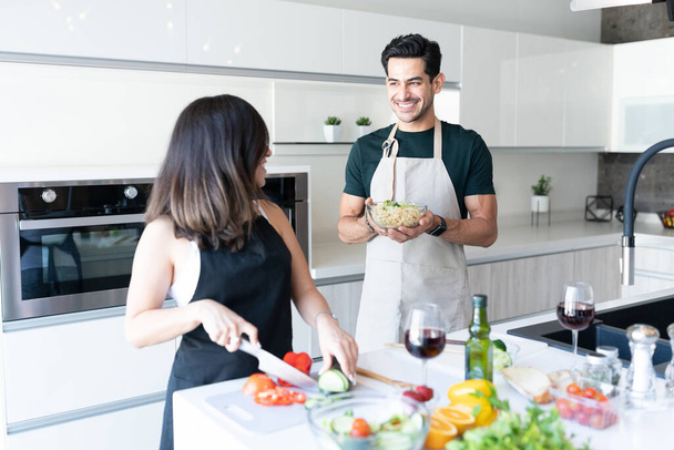 Sonriente joven sosteniendo un tazón de pasta mientras su novia corta verduras - Foto, imagen