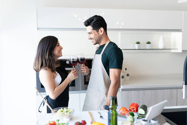 Sonriente joven pareja tostando gafas de vino mientras está de pie junto a la isla de la cocina - Foto, imagen