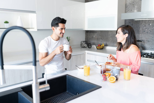 Smiling Hispanic young couple having breakfast at kitchen island - Foto, afbeelding