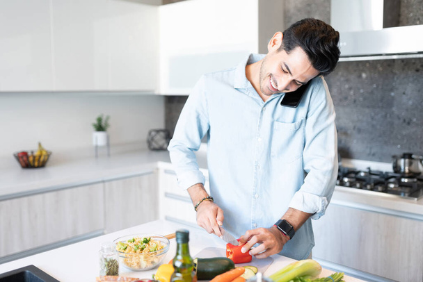 Lächelnder gutaussehender Mann spricht auf Smartphone, während er zu Hause Gemüse hackt - Foto, Bild