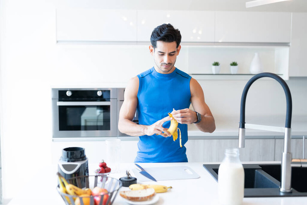 Junger athletischer Mann schält Banane, während er auf Kücheninsel steht - Foto, Bild