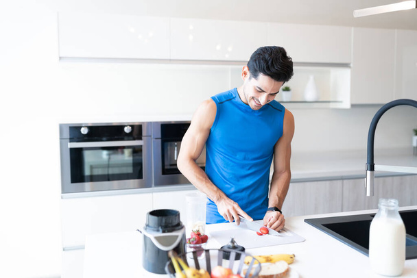 Latein schöner Mann schneidet Erdbeere auf Kücheninsel zu Hause in sportlichem Outfit - Foto, Bild
