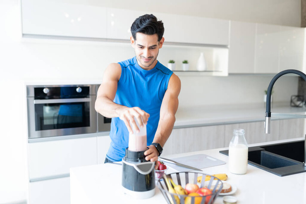 Добре виглядає активний чоловік готує полуничний молочний коктейль на кухні перед вправами
 - Фото, зображення