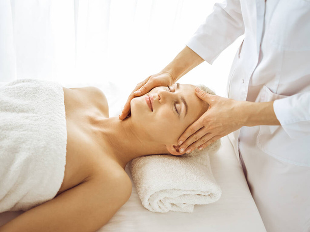 Beautiful blonde woman enjoying facial massage with closed eyes. Relaxing treatment in medicine and spa center concepts - Photo, image