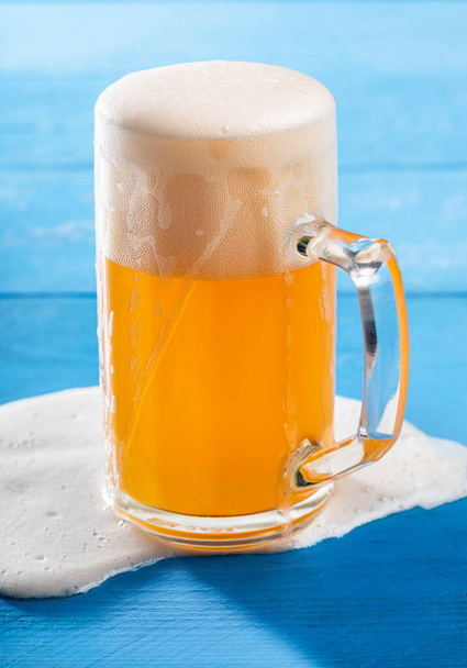 青い木製のテーブルの上にこぼれた泡でパイント内の濾過されていないビール。ブルーに隔離されたドイツの小麦ビールのピントのクローズアップ。夏の冷たい飲み物. - 写真・画像
