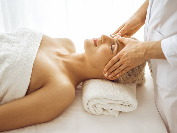 Beautiful blonde woman enjoying facial massage with closed eyes. Relaxing treatment in medicine and spa center concepts - Fotó, kép