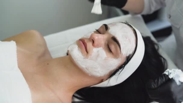 esteticista aplicación de peeling enzimático en las mujeres cara en el spa. Aplica una máscara blanca con un cepillo. Procedimiento cosmético en un moderno salón de belleza - Imágenes, Vídeo