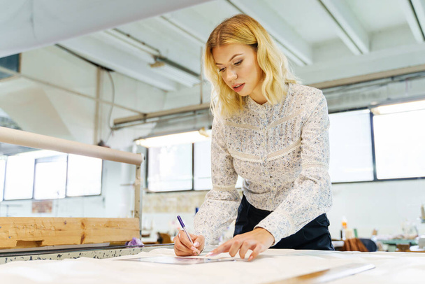 Молодая взрослая блондинка-кавказка в швейной мастерской, шьет новое дизайнерское эскизное платье на материале - Работающая за столом предпринимательница - Концепция гендерного равенства малого бизнеса - Фото, изображение