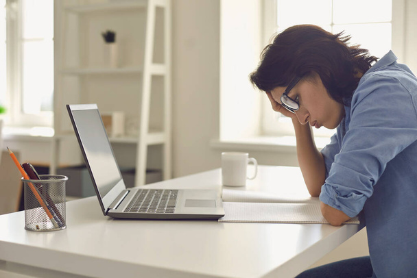 Уставшая молодая женщина, работающая над сложной задачей перед ноутбуком в домашнем офисе. Фрилансер чувствует усталость на работе - Фото, изображение
