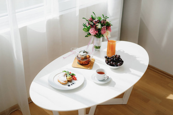White table inside the room near the window with a bouquet of flowers and breakfast in the morning before the wedding of tea, croissant and grapes with gold rings on a plate by the teapot. - Фото, изображение