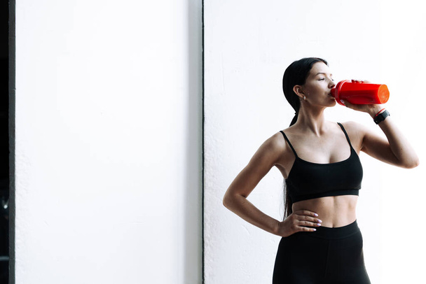 Horizontal photo of an Asian sportswoman in a black top and leggings who drinks water from a red bottle on a white background - Photo, Image