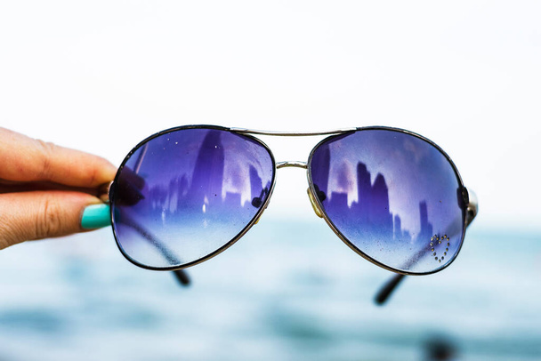 Female hand holds colorful sunglasses against the background of the sky and blue sea. The points reflect the storey buildings of the city. - Foto, imagen
