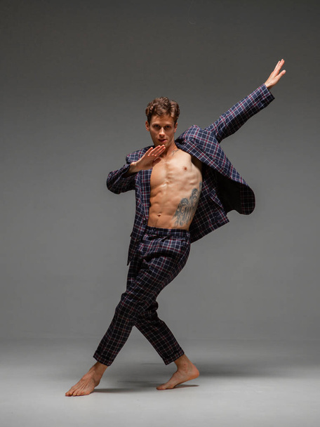 Stylish young guy dancer dancing in suit with naked torso isolated on gray background. Body with tattoo - 写真・画像