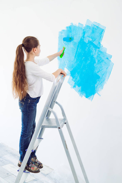 Επισκευή στο διαμέρισμα. Ευτυχισμένο κορίτσι ζωγραφίζει τον τοίχο με μπλε μπογιά, - Φωτογραφία, εικόνα