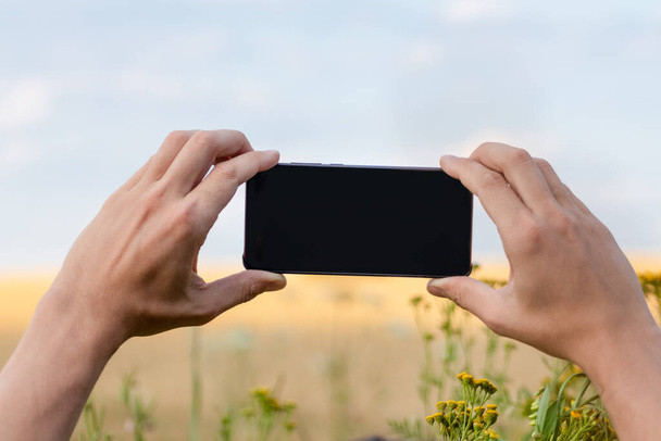 青い空の自然景観モックアップテンプレートと日当たりの良い花のフィールドに水平方向に両手で黒い画面とスマートフォンを保持 - 写真・画像